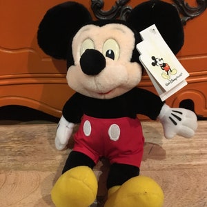 Może przedstawiać: Pluszowa zabawka Mickey Mouse siedząca na drewnianej powierzchni. Mickey ma na sobie swoje charakterystyczne czerwone spodenki z białymi guzikami i ż&oacute;łte buty. Ma czarne uszy i czarny nos. Zabawka ma białą metkę przymocowaną do prawego ucha, na kt&oacute;rej widnieje napis "Walt Disney World".