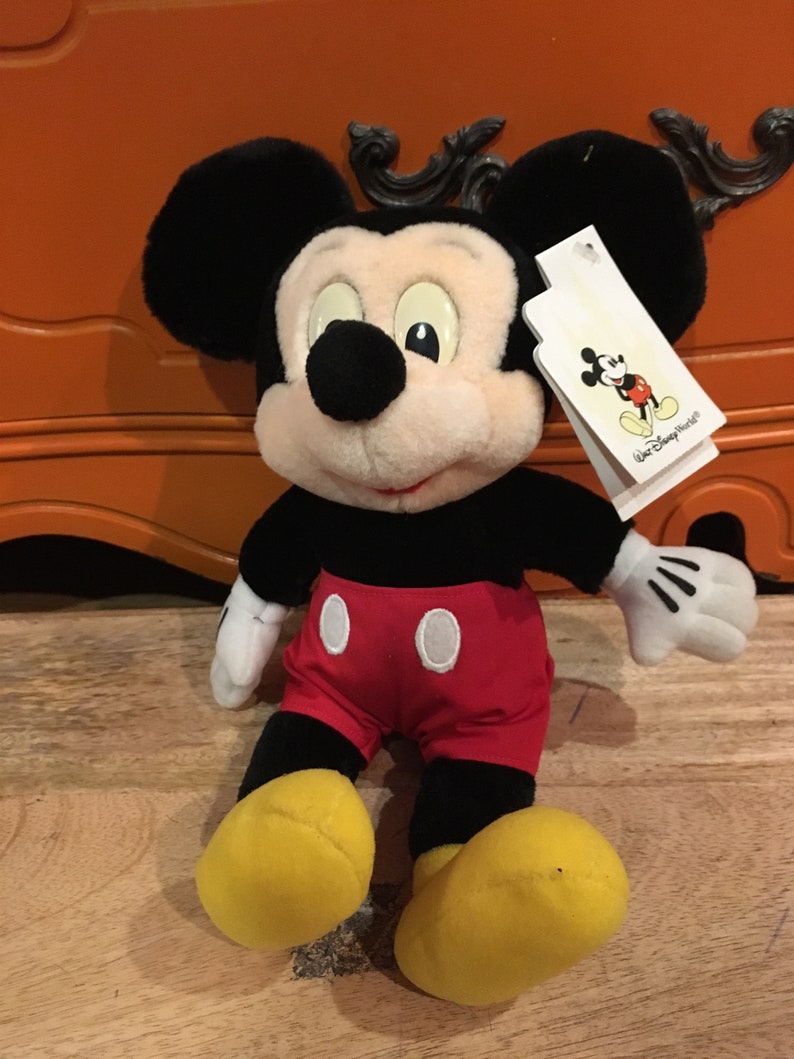 Może przedstawiać: Pluszowa zabawka Mickey Mouse siedząca na drewnianej powierzchni. Mickey ma na sobie swoje charakterystyczne czerwone spodenki z białymi guzikami i ż&oacute;łte buty. Ma czarne uszy i czarny nos. Zabawka ma białą metkę przymocowaną do prawego ucha, na kt&oacute;rej widnieje napis "Walt Disney World".