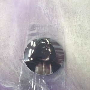 May include: A round, silver-colored pin with a black and white image of Darth Vader from Star Wars.