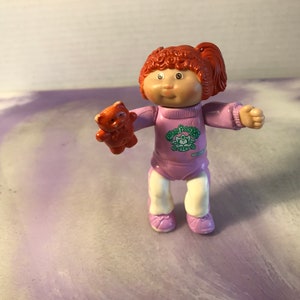 以下が含まれることがあります： 赤い髪の毛のキャベツパッチキッズ人形は、緑色のキャベツパッチキッズのロゴが入った紫色のロンパースを着ています。人形は小さなオレンジ色のぬいぐるみを持っています。