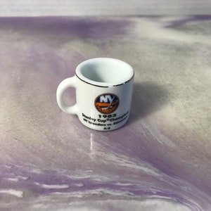 Może przedstawiać: Biały kubek ceramiczny z niebiesko-pomarańczowym logo New York Islanders. Kubek upamiętnia mistrzostwo Pucharu Stanleya z 1983 roku z tekstem "1983 Stanley Cup® Champions NY Islanders vs. Edmonton 4-0".