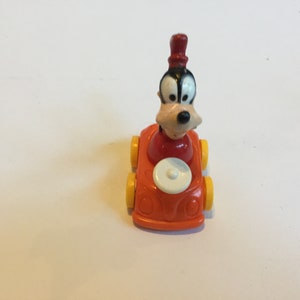 May include: A red and yellow toy car with a Goofy figure in the driver's seat. Goofy is wearing a red hat and has a black nose.