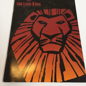 Puede incluir: Un programa negro y naranja para el musical de Broadway El Rey León. La portada presenta una imagen estilizada de la cara de un león.