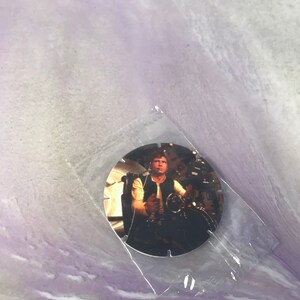Può includere: Una carta da collezione rotonda, argentata, con un'immagine ravvicinata di Han Solo di Star Wars. Han Solo indossa un gilet marrone ed è seduto nella cabina di pilotaggio del Millennium Falcon.