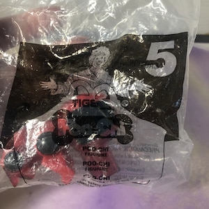 May include: A red plastic Poo-Chi figurine with black eyes and a black nose. The figurine is in a clear plastic bag with a Tiger logo and the number 5. The bag also has text that reads "Poo-Chi Figurine" and "Precaution".