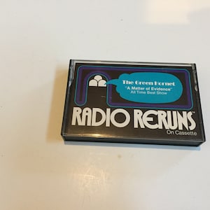 Puede incluir: Una cinta de casete negra con un gráfico colorido de un arco estilizado y el texto "The Green Hornet" "A Matter of Evidence" "All Time Best Show" RADIO RERUNS™ On Cassette.