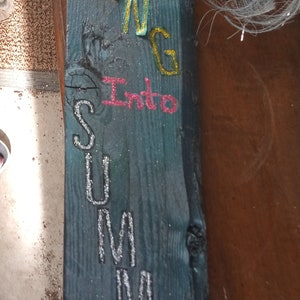 Puede incluir: Un letrero de madera rústico con las palabras "SPRING Into SUMMER" en letras brillantes. El letrero está decorado con flores artificiales y una piña dorada. El fondo es azul desgastado.