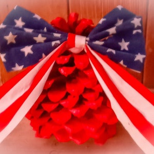 Puede incluir: Decoración patriótica con una base de piña roja y un lazo de tela. El lazo tiene un fondo azul con estrellas blancas y cintas a rayas rojas y blancas, evocando la bandera estadounidense.