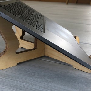 May include: A wooden laptop stand with a natural finish. The stand is designed to elevate a laptop, improving ergonomics. The laptop is a silver color. The stand has a curved design and is made of layered wood.