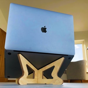May include: A light gray laptop with a black Apple logo rests on a natural wood laptop stand. The stand is made of interlocking pieces of light-colored wood, with a minimalist design. The laptop is angled upward.