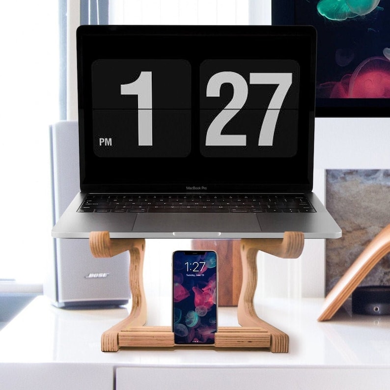 May include: A wooden laptop stand with a laptop and a smartphone on top. The laptop screen shows a digital clock displaying 1:27 PM. The smartphone screen shows a digital clock displaying 1:27. The stand is made of light brown wood and has a simple design.
