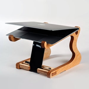 May include: A wooden laptop stand with a natural finish. The stand has a raised platform for a laptop and a lower shelf for a tablet or other items. The stand is designed to elevate the laptop to a more ergonomic viewing height.