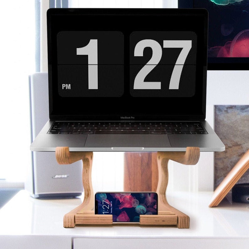 May include: A wooden laptop stand with a laptop on top and a smartphone in the stand's built-in phone holder. The laptop screen shows a digital clock displaying 1:27 PM.