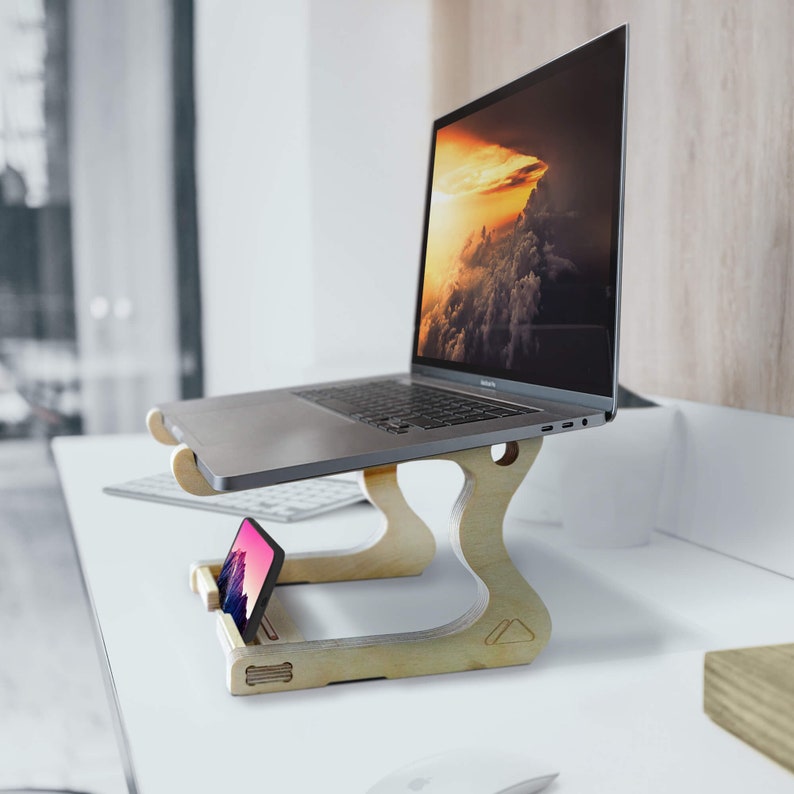 May include: A wooden laptop stand with a laptop and a smartphone on it. The laptop is open and has a screen displaying a landscape image of clouds and a sunset. The stand is made of light wood and has a simple design.