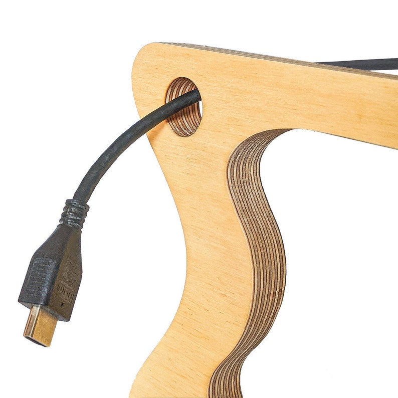 May include: A wooden cable organizer with a cutout for a cable. The organizer is made of light wood and has a natural finish. A black cable is threaded through the cutout.