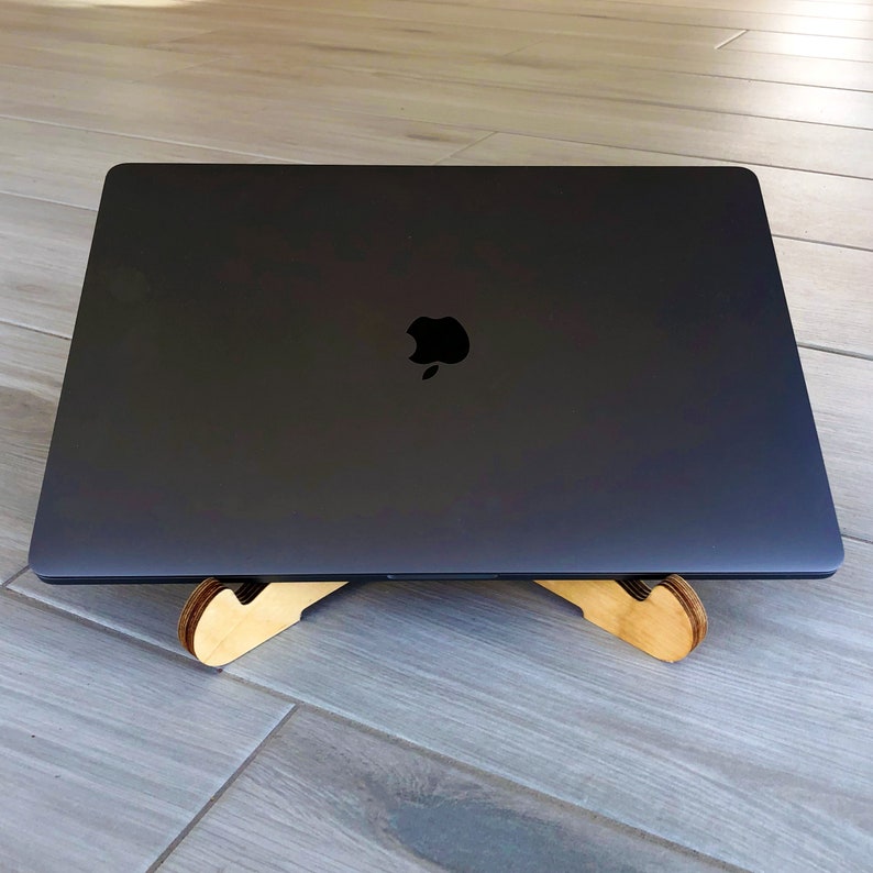 May include: A closed, dark gray laptop with the Apple logo is placed on a light-colored wooden laptop stand. The stand has a curved design and is made of light-colored wood. The laptop is positioned on a light gray wooden floor.