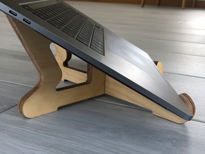 May include: A wooden laptop stand with a natural finish. The stand is designed to elevate a laptop, improving ergonomics. The laptop is a silver color. The stand has a curved design and is made of layered wood.