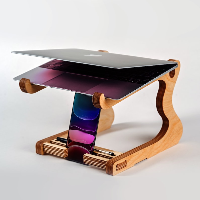 May include: A wooden laptop stand with a laptop on top and a phone in a stand on the side. The stand has a slot for a pen or pencil.
