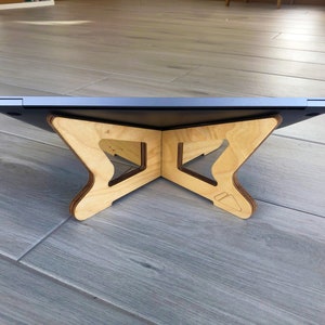 May include: A wooden laptop stand with a natural finish. The stand has a unique, geometric design with a cross-shaped base and angled supports. The laptop sits on top, creating an ergonomic workspace. The stand is designed to elevate the laptop for better viewing.