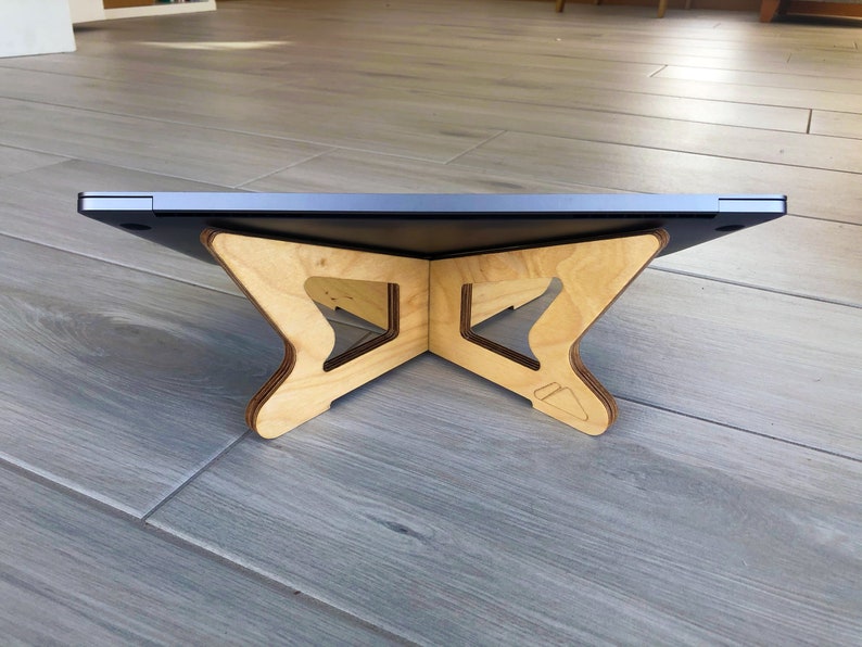 May include: A wooden laptop stand with a natural finish. The stand has a unique, geometric design with a cross-shaped base and angled supports. The laptop sits on top, creating an ergonomic workspace. The stand is designed to elevate the laptop for better viewing.