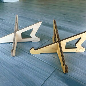 May include: Two wooden display stands with a natural wood finish. Each stand has a unique cutout design and a curved base. The stands are designed to hold items and are made of layered wood.