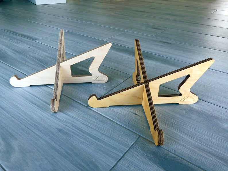 May include: Two wooden display stands with a natural wood finish. Each stand has a unique cutout design and a curved base. The stands are designed to hold items and are made of layered wood.