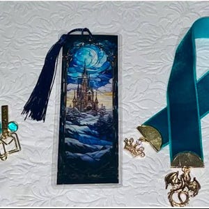 May include: Three bookmarks with decorative charms. One features a castle scene under a full moon, another is a teal ribbon with a dragon charm, and the third has a book charm with a teal tassel. All are on a white textured surface.