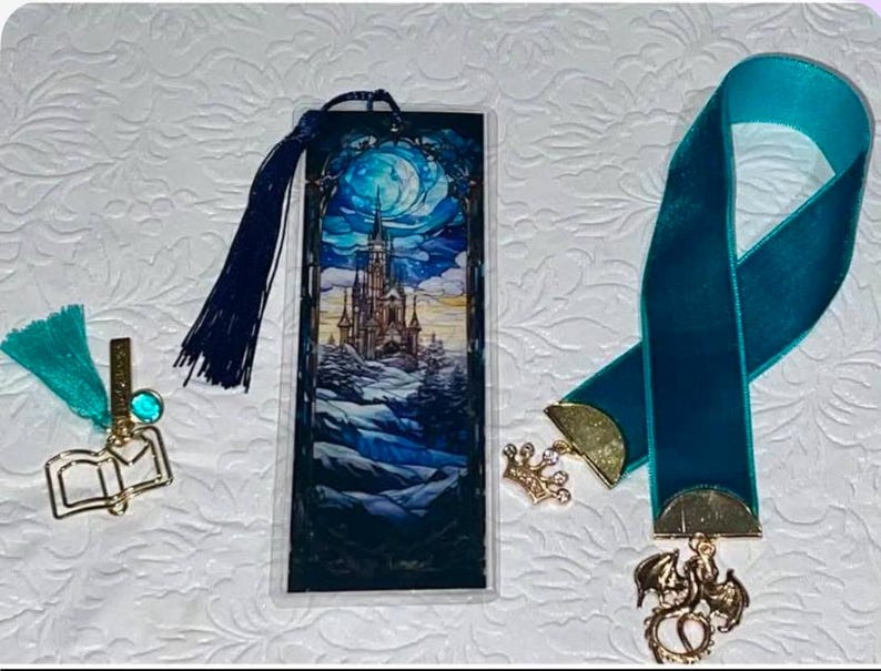 May include: Three bookmarks with decorative charms. One features a castle scene under a full moon, another is a teal ribbon with a dragon charm, and the third has a book charm with a teal tassel. All are on a white textured surface.