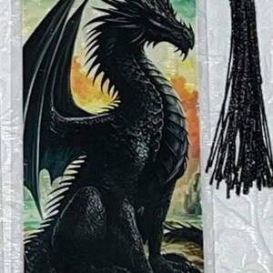 May include: A fantasy-themed bookmark featuring a detailed illustration of a black dragon with large wings, set against a colorful background of a castle and sunset. The bookmark has a black tassel.