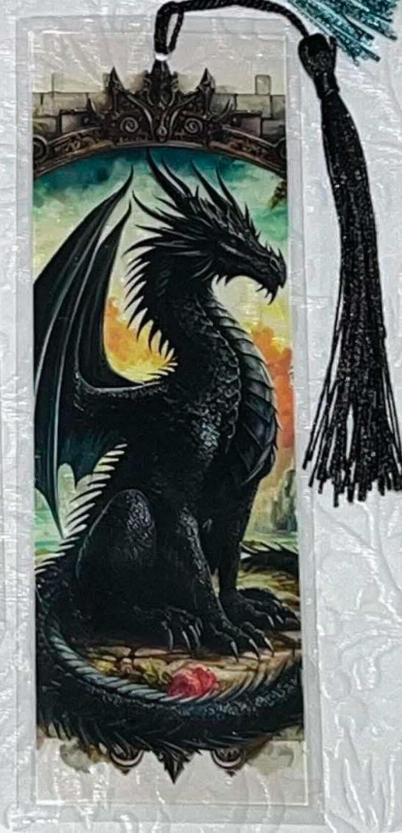 May include: A fantasy-themed bookmark featuring a detailed illustration of a black dragon with large wings, set against a colorful background of a castle and sunset. The bookmark has a black tassel.