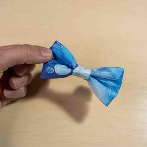 May include: A blue bow tie with a white and blue pattern. The bow tie is made of fabric and has a classic bow shape. The fabric has a watercolor-like design with shades of blue and white.