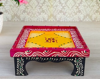 May include: A decorative wooden platform, rectangular in shape, featuring a yellow top with a central symbol, framed by a red border with white and pink patterns. The black legs are decorated with white and green floral designs.