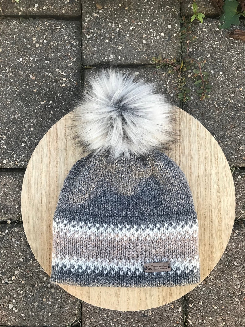 Puede incluir: Un gorro de punto gris y blanco con una etiqueta de cuero marr&oacute;n que dice "Made in Canada". El gorro tiene un pomp&oacute;n de piel sint&eacute;tica blanca en la parte superior.