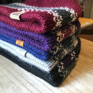 Puede incluir: Una pila de cuatro gorros de punto de diferentes colores y patrones. Los gorros est&aacute;n hechos de un hilo suave y c&aacute;lido y tienen una sensaci&oacute;n acogedora. El gorro de arriba es burdeos con rayas blancas y grises, el segundo gorro es morado, el tercer gorro es azul y el gorro de abajo es gris con rayas blancas y moradas.