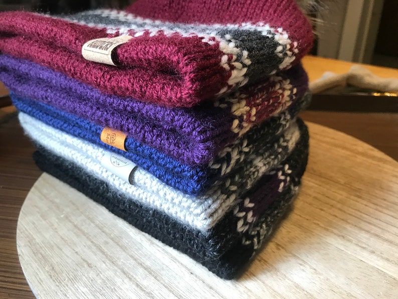 Puede incluir: Una pila de cuatro gorros de punto de diferentes colores y patrones. Los gorros est&aacute;n hechos de un hilo suave y c&aacute;lido y tienen una sensaci&oacute;n acogedora. El gorro de arriba es burdeos con rayas blancas y grises, el segundo gorro es morado, el tercer gorro es azul y el gorro de abajo es gris con rayas blancas y moradas.