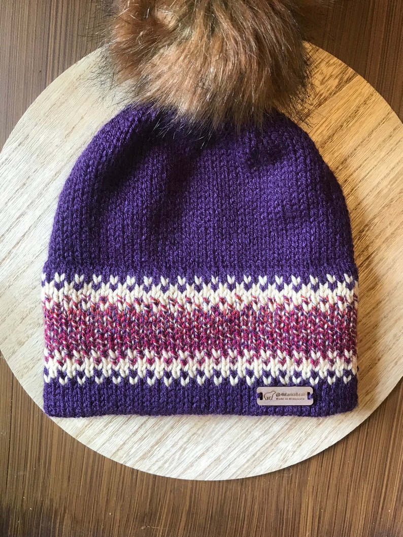 Puede incluir: Gorro de punto morado con una banda estampada en blanco y rosa y un pomp&oacute;n de piel sint&eacute;tica marr&oacute;n. El gorro tiene una etiqueta que dice "Hecho a mano en Minnesota".