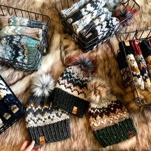 Könnte beinhalten: Eine Sammlung von Strickmützen mit Bommeln in verschiedenen Farben und Mustern, darunter Schwarz, Weiß, Blau, Grün, Braun und Orange. Die Mützen werden in Drahtkörben auf einem Kunstfell präsentiert.