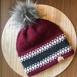 Puede incluir: Un gorro de punto burdeos con un patr&oacute;n de rayas blancas y grises y un pomp&oacute;n de piel sint&eacute;tica gris. El gorro tiene un parche de cuero marr&oacute;n con el texto "CENTRAL RAIDERS".