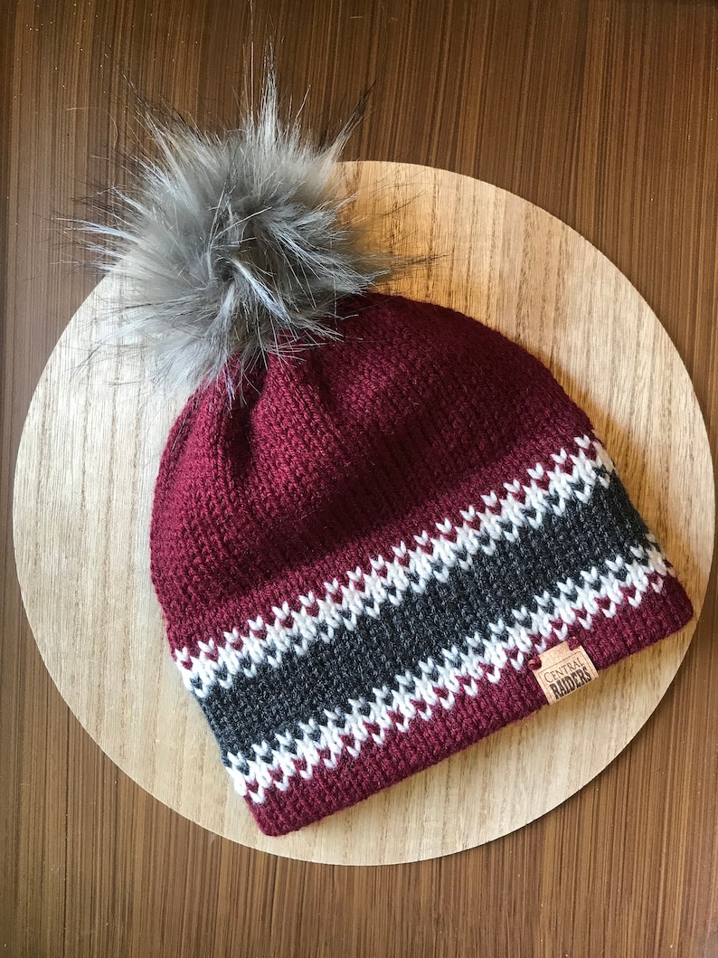 Puede incluir: Un gorro de punto burdeos con un patr&oacute;n de rayas blancas y grises y un pomp&oacute;n de piel sint&eacute;tica gris. El gorro tiene un parche de cuero marr&oacute;n con el texto "CENTRAL RAIDERS".