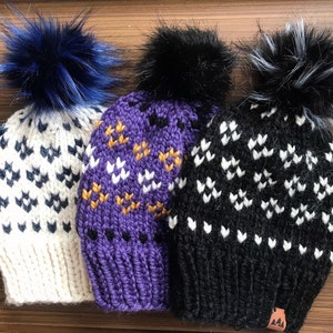 Puede incluir: Tres gorros de invierno de punto con pompones. Los gorros son blancos con patrones geométricos negros y azules, morados con patrones geométricos dorados y blancos, y negros con patrones geométricos blancos.