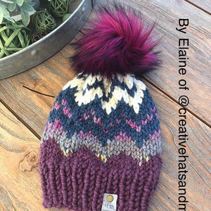 May include: A handmade knit hat with a large, fluffy, magenta pom-pom. The hat features a chevron pattern in shades of purple, blue, cream, and pink. The label reads "cfm creative hats and more". The text "By Elaine of @creativehatsandmore" is also visible.