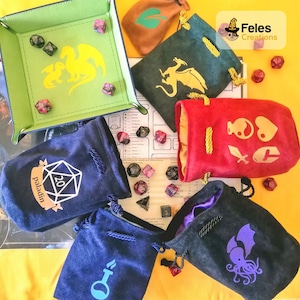 May include: A collection of six velvet dice bags with various designs. The bags are in different colours, including red, green, blue, and black. The designs include a dragon, a sword, a heart, a yin-yang symbol, a key, and an octopus. The bags are on a yellow surface with dice scattered around them.