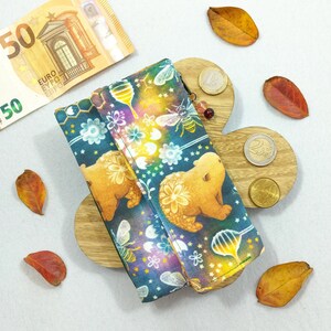 Può includere: Un portafoglio colorato con un motivo a orsi, api e favi. Il portafoglio è aperto, rivelando l'interno. Banconote e monete in euro sono posizionate vicino, insieme a foglie autunnali.
