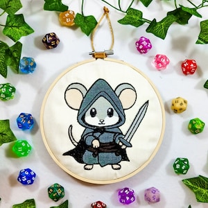 Può includere: Ricamo su cerchio raffigurante un topo dei cartoni animati vestito con un costume fantasy, che tiene una spada. Il topo indossa un mantello con cappuccio grigio e blu. L'opera d'arte è ambientata su uno sfondo bianco, circondata da dadi poliedrici colorati e edera verde.