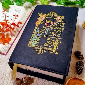 Op de afbeelding: Een marineblauwe boekomslag met de woorden "Once Upon a Time" geborduurd in goud, roze en blauw. De omslag heeft een sierlijk ontwerp met vlinders en bloemmotieven. Een gouden lint-bladwijzer is zichtbaar.