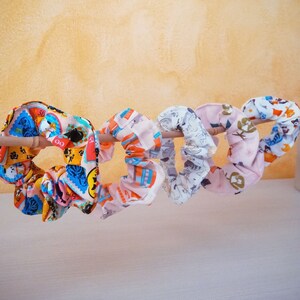 Può includere: Cinque scrunchies in tessuto colorati con vari motivi, tra cui gatti, fiori e disegni geometrici. Gli scrunchies sono appesi a un bastoncino di legno.