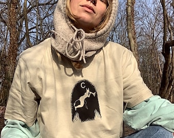May include: A person wearing a beige t-shirt with a black and white graphic of a figure holding a horn. The t-shirt has a crescent moon above the figure. The person is wearing a light beige knit hooded scarf.