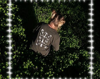 May include: A person wearing a dark gray long-sleeve shirt with a white graphic print of a tree and other nature-inspired symbols. The shirt is worn in a lush green outdoor setting.