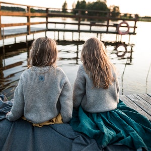 May include: Two children wearing gray sweaters and sitting on a gray blanket on a wooden dock. The child on the left is wearing a yellow skirt and the child on the right is wearing a teal skirt.