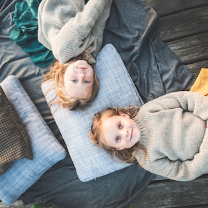 May include: Two children laying on a gray blanket with pillows. Both children are wearing gray sweaters and have long blonde hair. One child is wearing a green skirt and the other is wearing a yellow skirt.